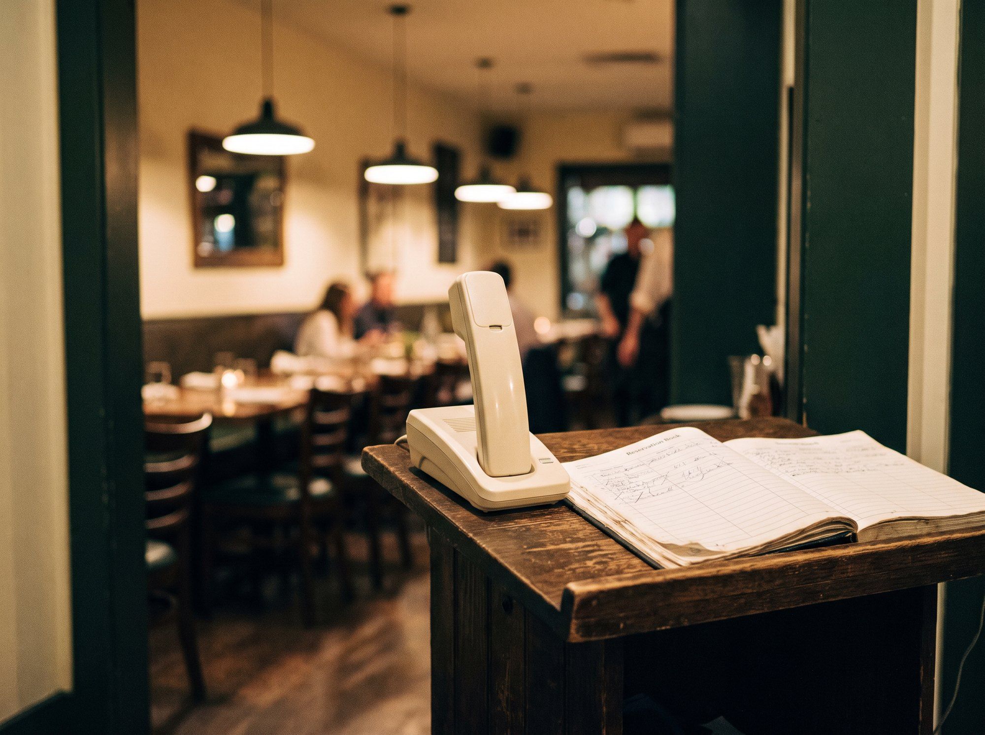 A phone and an open reservation book on a host stand, the dining room busy behind.